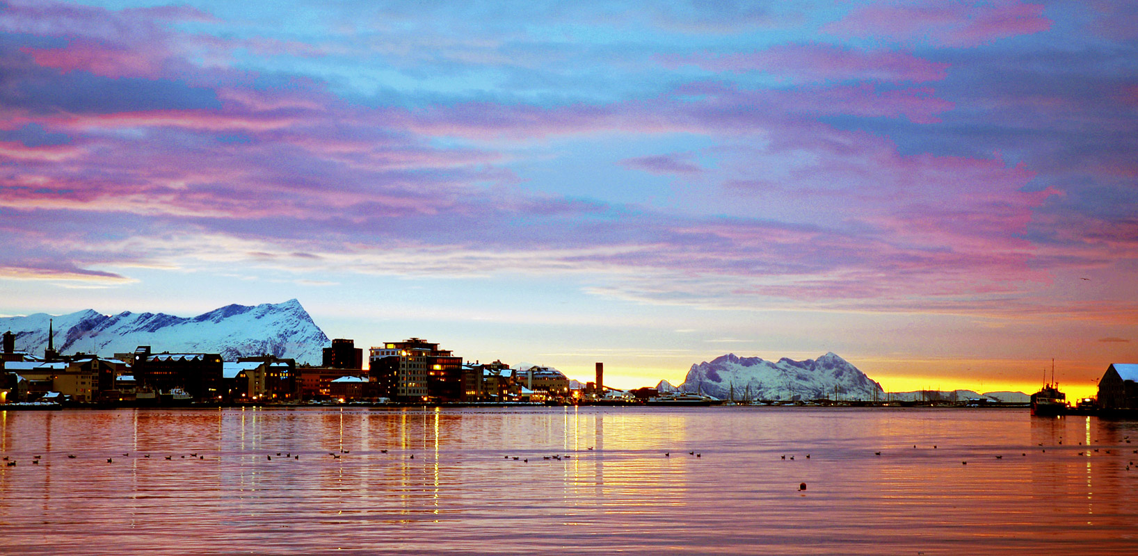 Bodø waterfront seen across the harbour at dawn in winter, snow-covered mountains behind the town and a fishing boat at the quay