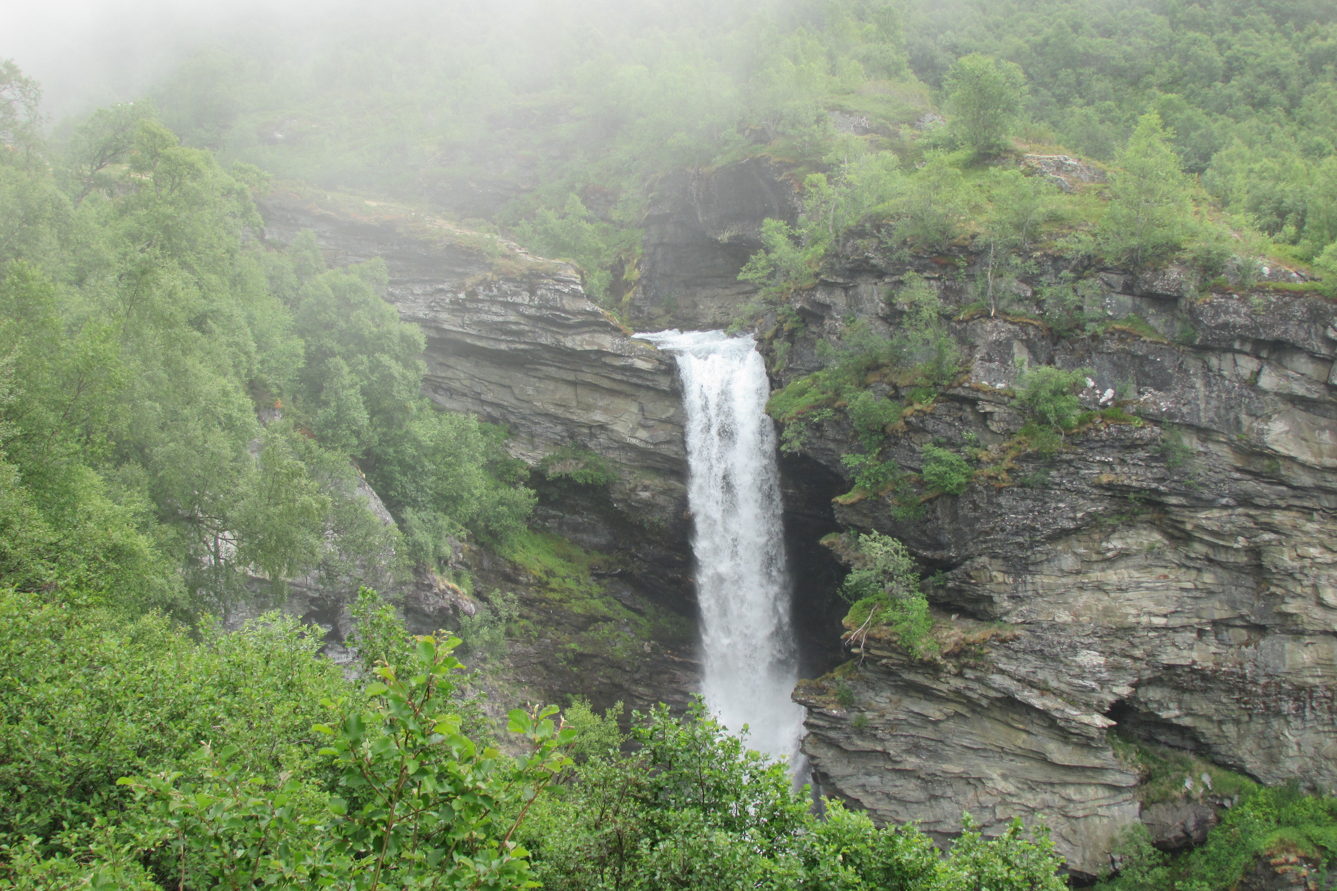 Storseterfossen waterfall dropping 30 meters from a dark rock overhang into mist above Geirangerfjord, Norway