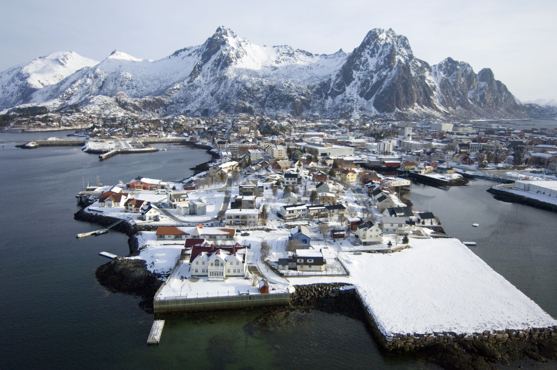 Aerial view of a snow-covered Lofoten harbour town on a low peninsula, with jagged white peaks behind and dark sea on both sides