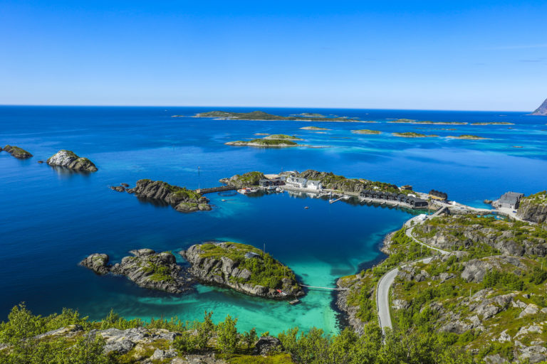A narrow road winding past a cluster of houses on small rocky islets joined by bridges, with turquoise water and the open sea behind, on the outer coast of Senja in summer